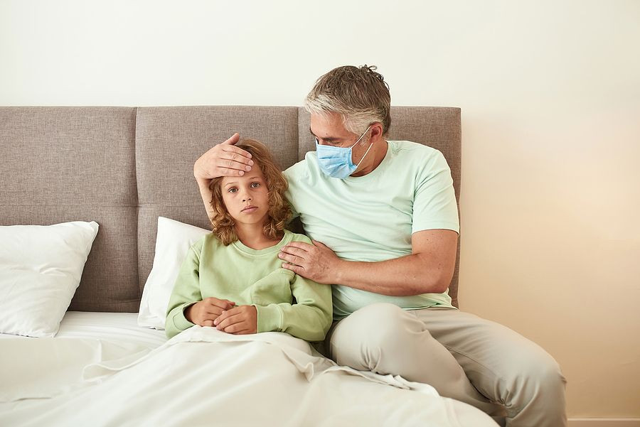 Portrait Of Loving Father In Facemask Take Care Of Sick Unwell Teen Son Stay In Bed Suffer From Covi