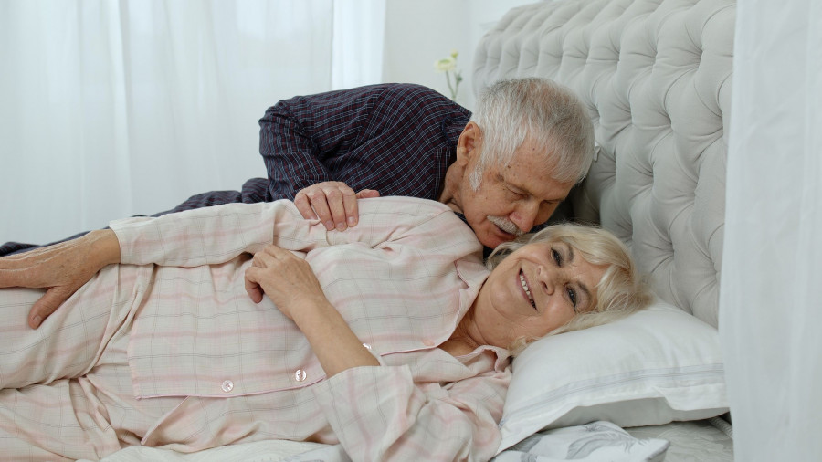 Senior Caucasian Grandparents Couple Lying In Bed At Home In Morning. Man Gently Whispering To Woman